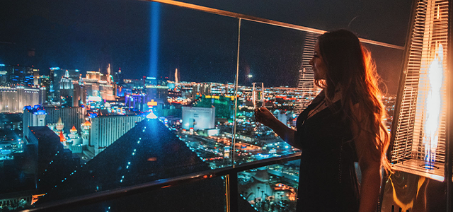 Woman looking out the window of Skyfall Lounge at Delano Rooftop bar with a drink in hand; Luxor hotel lit up